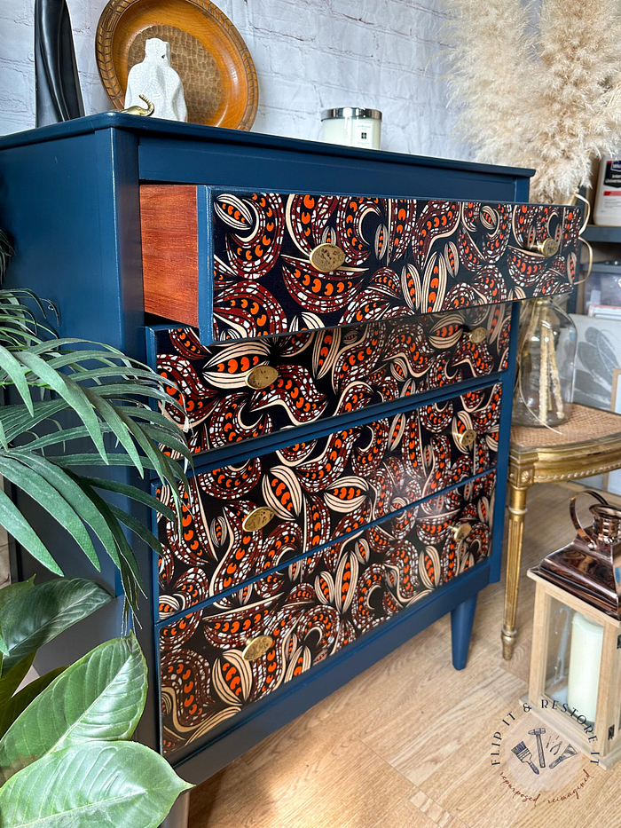 A blue dresser with four drawers, featuring intricate mixed brown, orange, and white paisley patterns. The top drawer is slightly pulled out. The dresser is placed near a green plant and various home decor items. The floor has a circular design.