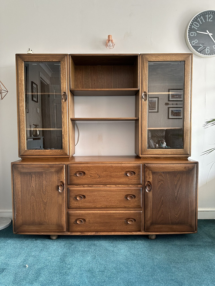 The Ercol Windsor Sideboard and Display Cabinets with Lighting in Golden Dawn Finish stands on a blue carpet. It features glass-paneled doors on the upper sections and solid doors on the lower sections, along with three central drawers and shelves in the middle. To the right, a wall clock is visible, and framed pictures adorn the wall.