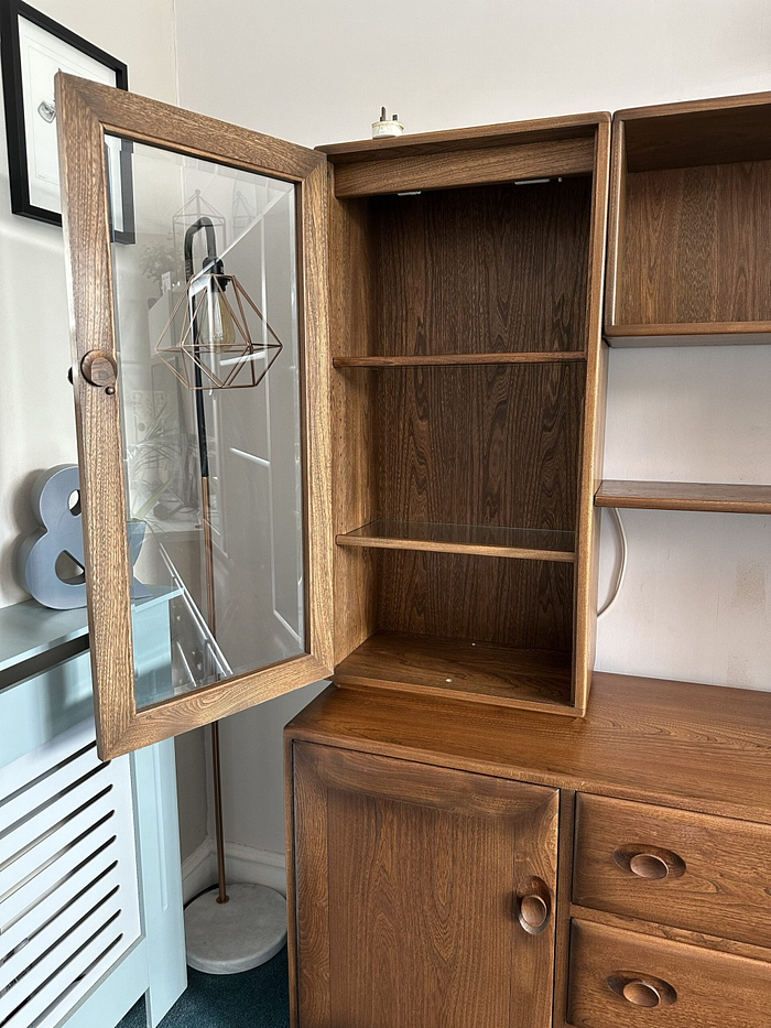 The Ercol Windsor Sideboard and Display Cabinets with Lighting in Golden Dawn Finish features a glass door, revealing empty shelves inside. Below the shelving area, the cabinet includes drawers and a wooden-door storage compartment. Positioned against a wall in a room, it stands next to a radiator cover and a standing lamp.