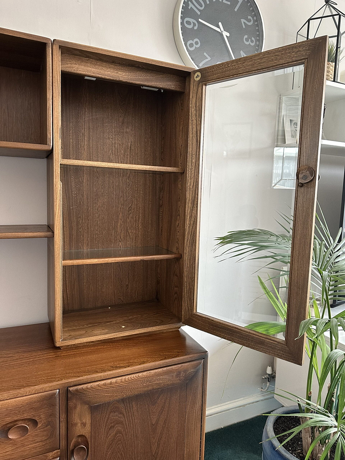 The Ercol Windsor Sideboard and Display Cabinet with Lighting in Golden Dawn Finish showcases two empty shelves behind an open glass door. The cabinet, featuring a polished finish, is set against a wall adorned with a clock, plant, and glass lantern in the backdrop.