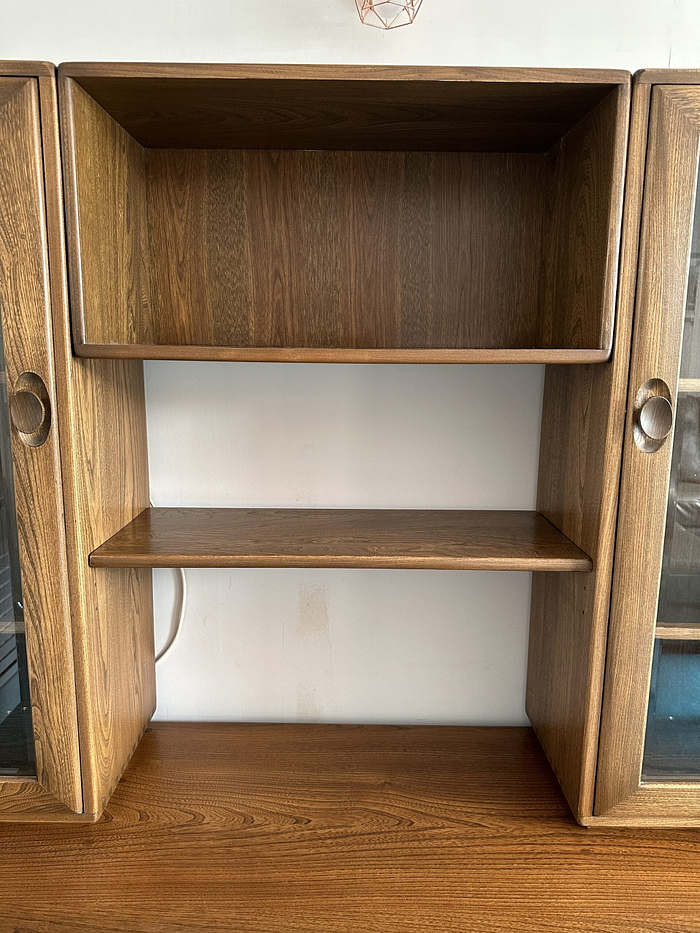 The Ercol Windsor Sideboard and Display Cabinets with Lighting in a Golden Dawn finish features two glass-paneled doors on either side and an open middle section. The middle section includes a wide shelf halfway up and another enclosed space above it. The cabinet rests against a white wall, with a visible power cord behind it.