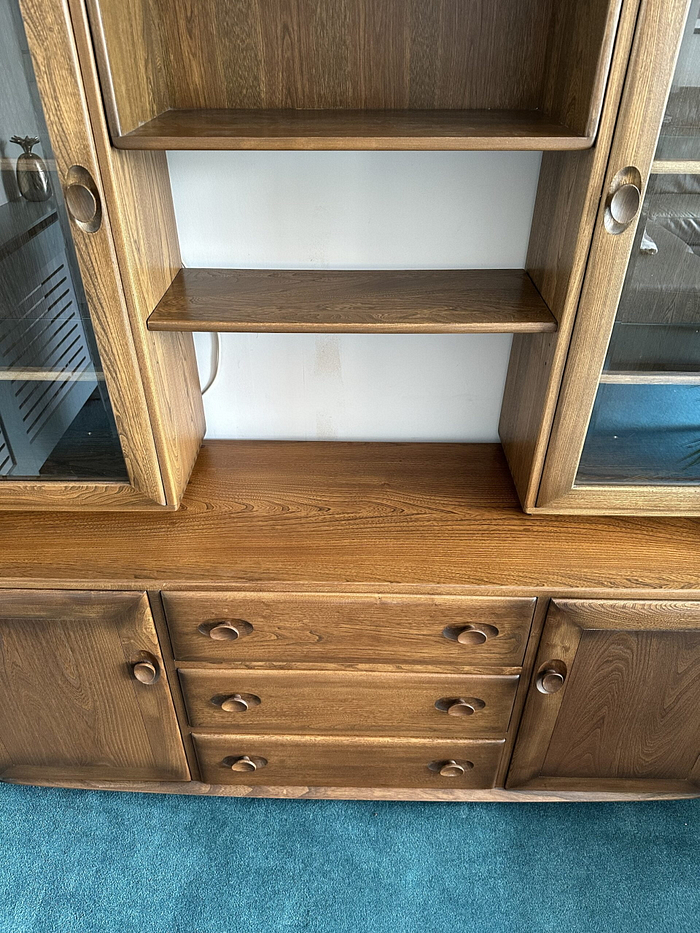 The Ercol Windsor Sideboard and Display Cabinets with Lighting in Golden Dawn Finish is showcased. It features a central section with three drawers beneath two open shelves, flanked by glass-fronted display sections on either side. The cabinet is set against a white wall, and the floor is covered with blue carpet.