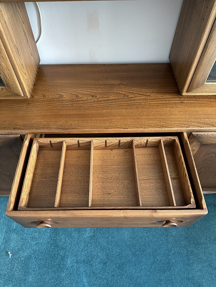 The Ercol Windsor Sideboard and Display Cabinets with Lighting in Golden Dawn Finish feature an open wooden drawer, revealing several long, vertical compartments inside. This functional piece is part of a larger furniture unit, resting elegantly on a blue carpeted floor. The entire unit boasts a rustic appearance with a polished finish.