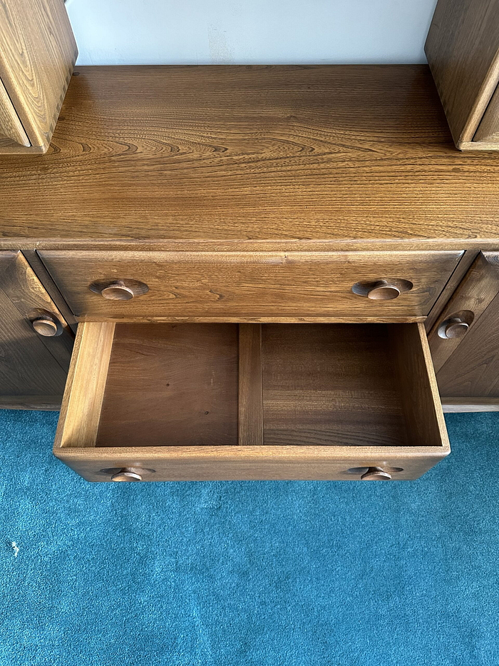 The Ercol Windsor Sideboard and Display Cabinet with Lighting in a Golden Dawn finish showcases its polished surface with visible wood grain. With an open drawer revealing two compartments, it features round wooden knobs and rests on a blue carpet.