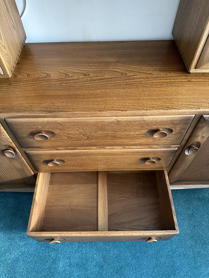 An Ercol Windsor Sideboard and Display Cabinet with a Golden Dawn finish, featuring multiple drawers and distinctive circular handles, stands on a blue carpeted floor. One drawer is open, revealing its empty interior divided by a single central divider.