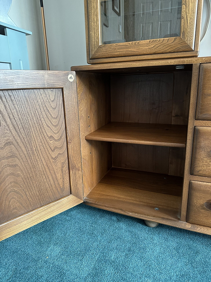 The Ercol Windsor Sideboard and Display Cabinet with Lighting in Golden Dawn Finish features a wooden structure with an open door, revealing two interior shelves. The cabinet is mounted with a mirror on top and is placed on a blue carpeted floor. Drawer handles are visible on the right side of the cabinet, and the interior appears empty.