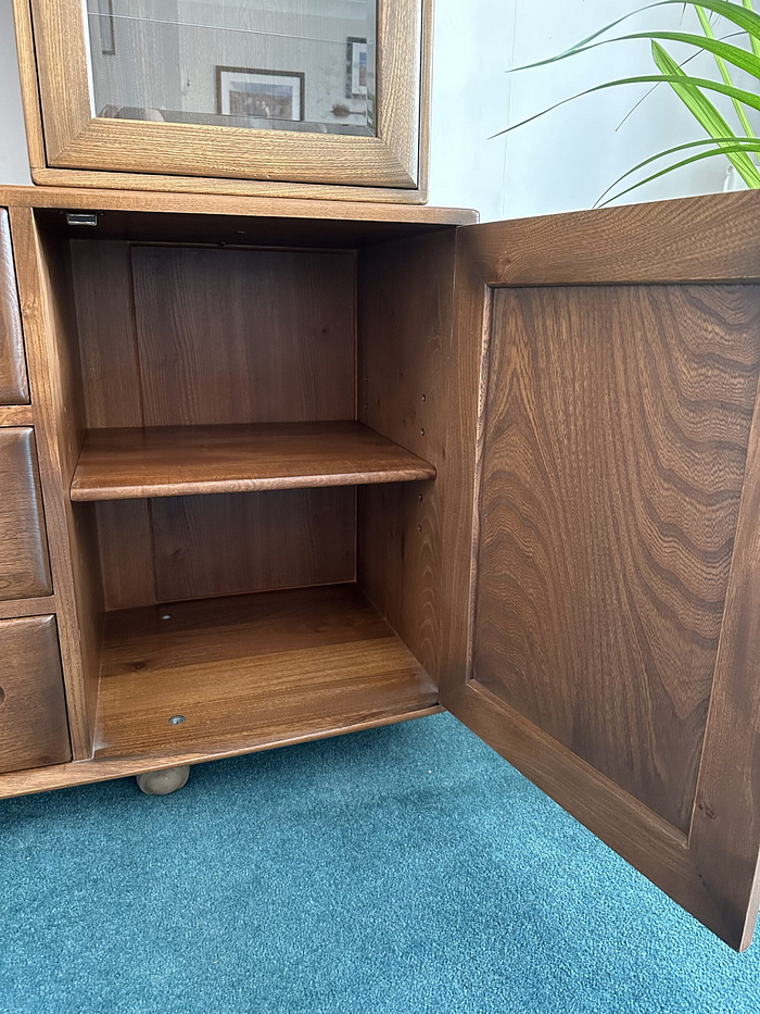 An Ercol Windsor Sideboard and Display Cabinet with Lighting, Golden Dawn Finish, with an open door revealing an empty interior shelf. The cabinet is situated on a blue carpet, and a green potted plant is partially visible in the background. The cabinet features a dark brown finish with natural wood grain visible.