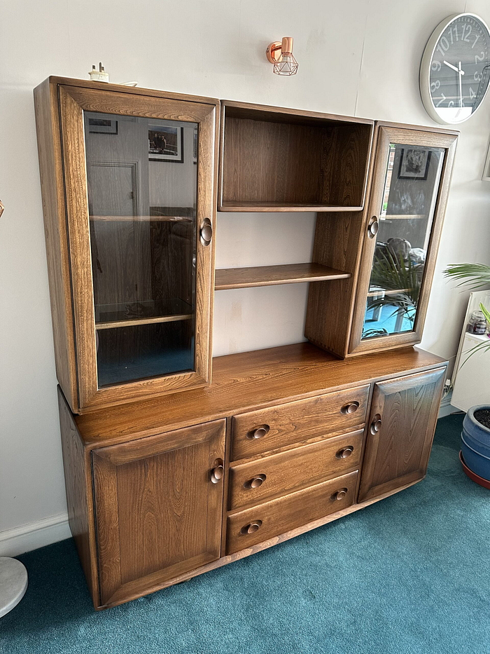 The Ercol Windsor Sideboard And Display Cabinets With Lighting in Golden Dawn Finish features glass doors and shelves on both sides, open compartments in the middle, and drawers and cabinets below. The sideboard stands on a teal carpeted floor with a wall clock visible in the background.