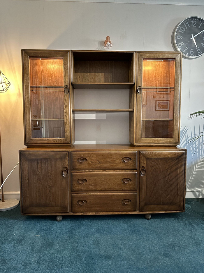 The Ercol Windsor Sideboard and Display Cabinets with Lighting in Golden Dawn Finish feature a mid-century modern design, combining open shelves and glass-fronted cabinets on the top with drawers and closed cabinets on the bottom. It is placed on a teal carpet, with a floor lamp and a wall clock visible in the background.