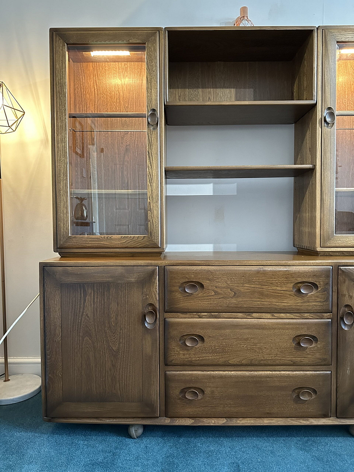 The Ercol Windsor Sideboard and Display Cabinets with Lighting in Golden Dawn Finish stand against the wall, showcasing glass-paneled doors on the upper left and right sides, with shelves centered in between. Below these, there are drawers and additional cabinet space. A floor lamp with a geometric design is positioned to the left. The floor features a blue carpet.