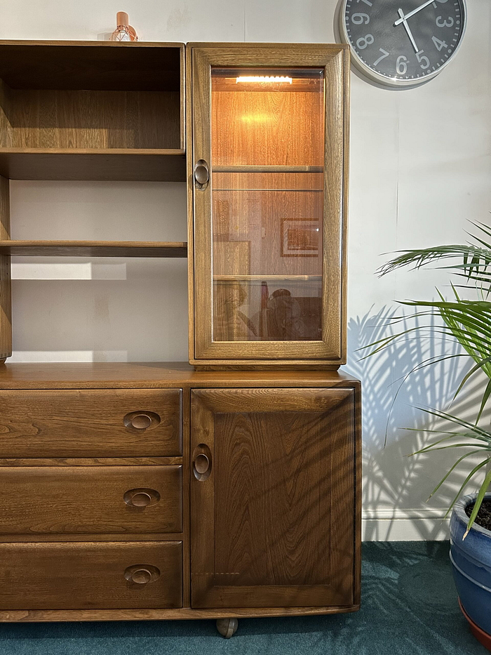 The Ercol Windsor Sideboard And Display Cabinets With Lighting in a Golden Dawn finish features a combination of drawers and shelves. On the right side, the upper section has a glass door with an illuminated interior, while the lower section has a wooden door. A wall clock is mounted above it, and a potted plant is partially visible on the right.