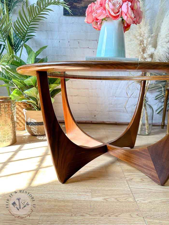 A stylish G-Plan Astro Round Coffee Table Mid-Century Vintage with a geometric base stands on a wooden floor. On the table is a teal vase with a bouquet of pink flowers. The scene is enhanced by the surrounding houseplants and the light streaming through a nearby window.