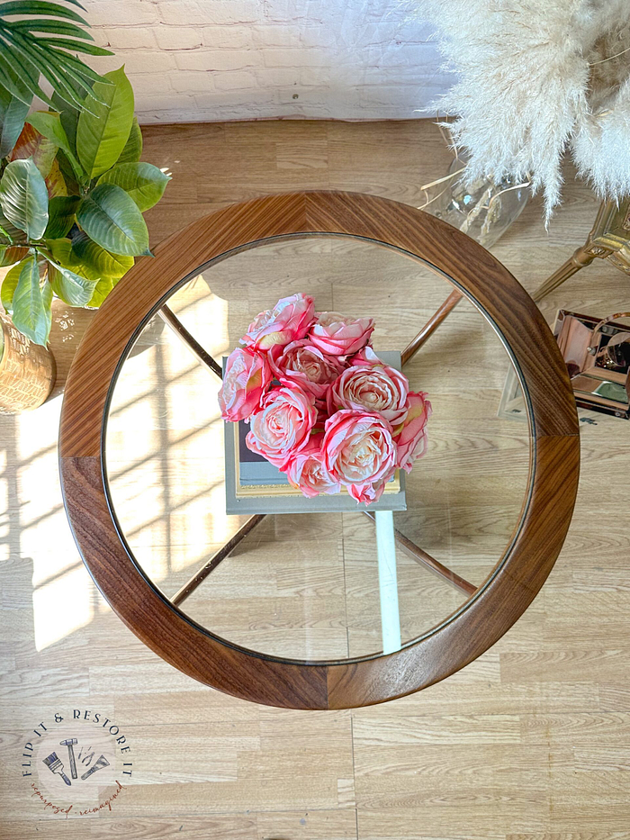 A G-Plan Astro Round Coffee Table Mid-Century Vintage, featuring a wooden frame and a glass top, holds a small bouquet of pink and white roses. The table is positioned on a wooden floor next to a green potted plant and a fluffy beige rug. Sunlight streams in from a nearby window, casting shadows on the floor.