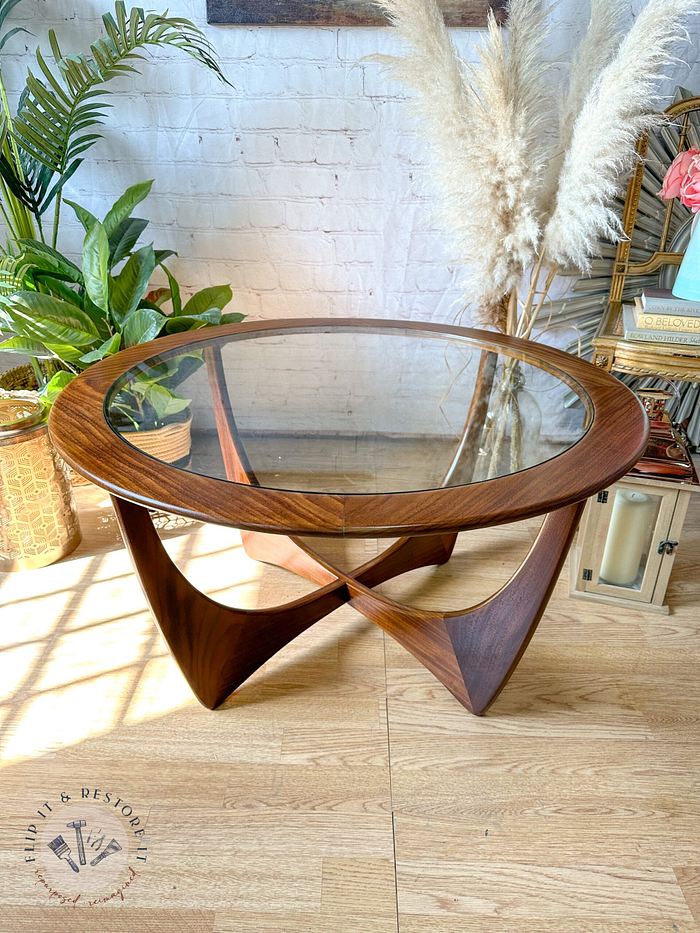 A G-Plan Astro Round Coffee Table Mid-Century Vintage with a sculpted wooden base sits on a hardwood floor. The background features a potted green plant, decorative pampas grass, and a wicker lamp beside a stack of books. The logo "Flip It & Restore It" is visible at the bottom left.