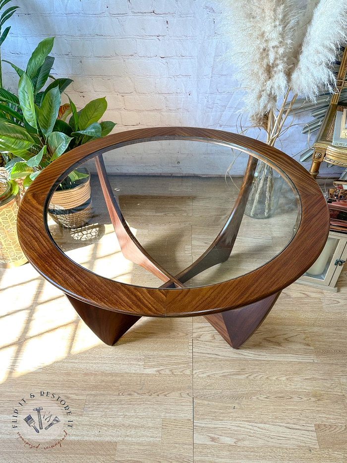 A G-Plan Astro Round Coffee Table Mid-Century Vintage with a wooden base sits on a hardwood floor. Behind it are some green leafy plants on the left and tall beige dried pampas grass on the right. There is a small shelf with miniature decorative items to the right of the table.