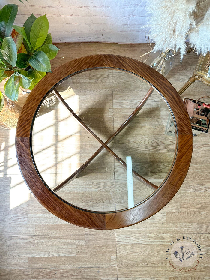 An overhead view of a G-Plan Astro Round Coffee Table Mid-Century Vintage on a wooden floor. The table features a circular wooden frame and cross-shaped wooden supports underneath the glass top. Beside the table, there is a potted plant with large green leaves, and a fluffy rug is nearby.