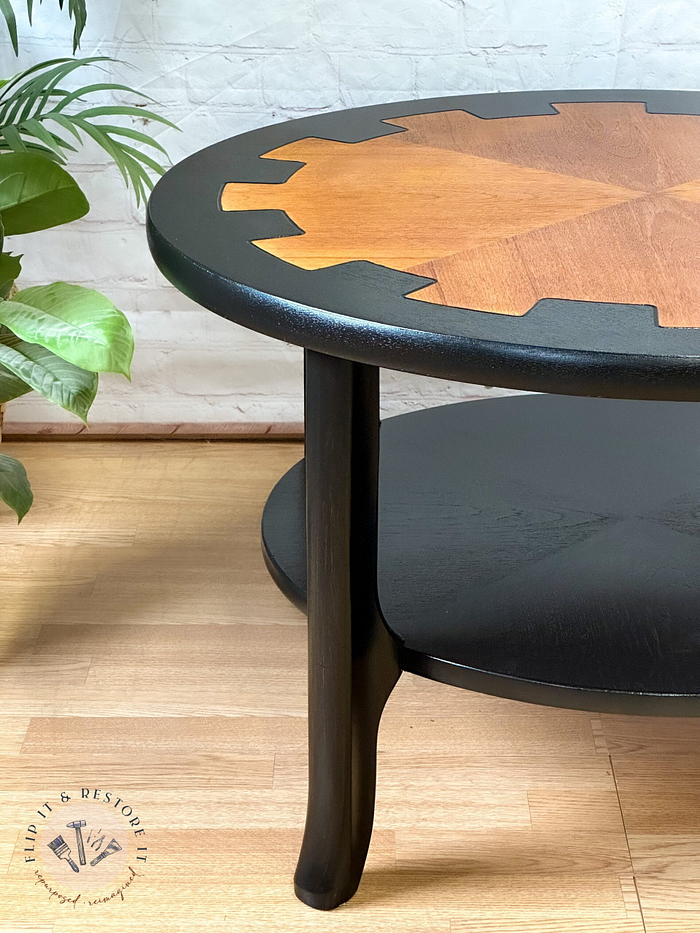 Here is a close-up of the MCM Circular Round 'Cog' Coffee Table by Nathan Furniture. This two-tier wooden side table features a top surface with a stained wood inlay showcasing geometric shapes, while the table legs and bottom tier are hand-painted in black. A green plant and light-colored flooring can be seen in the background.