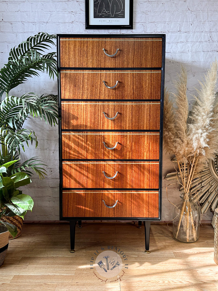 The Exquisite Mid Century MCM Walnut Tallboy 6 Drawers Chest Of Drawers by Meredew stands against a white brick wall, with its six drawers featuring curved metal handles. Lush green plants flank it on the left, while dried pampas grass in a clear vase is placed on the right. A framed minimalist artwork hangs above the dresser.