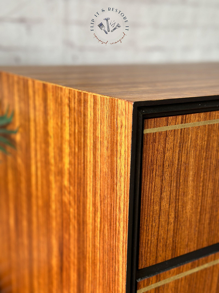 Close-up of the Exquisite Mid Century MCM Walnut Tallboy 6 Drawers Chest Of Drawers by Meredew, showcasing its smooth, lacquered finish. The corner of the chest is prominently featured, highlighting its precise edges and rich wood grain. Above the furniture is a circular logo with text encircling a paintbrush and hammer.