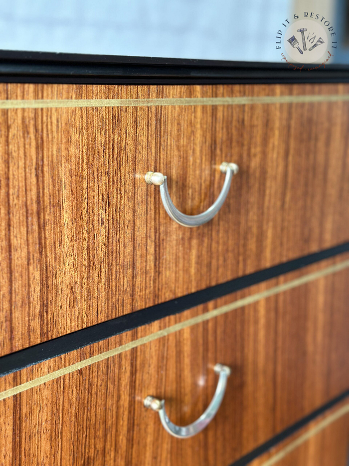 Close-up of an "Exquisite Mid Century MCM Walnut Tallboy 6 Drawers Chest Of Drawers by Meredew" featuring six drawers, each adorned with metallic handles. The wood grain runs horizontally, accented by thin black and gold inlays between the drawers. The top edge is finished in black, and the background displays a segment of a "Flip It & Restore It" logo.