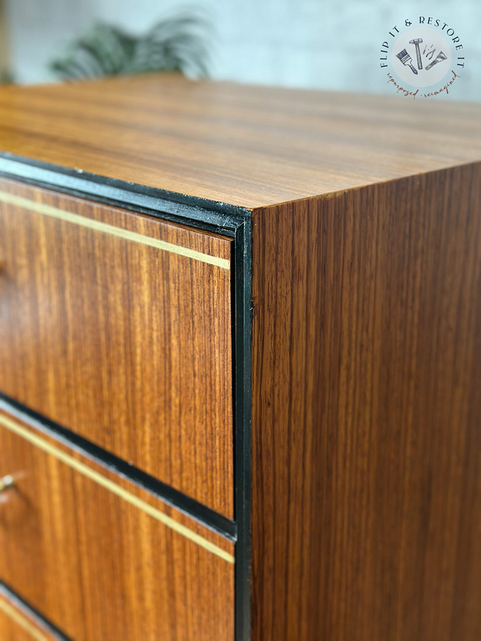 A close-up view of the Exquisite Mid Century MCM Walnut Tallboy 6 Drawers Chest Of Drawers by Meredew showcases a sleek, smooth finish with minimalist design elements. The tallboy features subtle beige lines as decorative accents and a polished surface. In the background, part of a plant is visible. The logo in the top-right corner says "Flip It & Restore It.