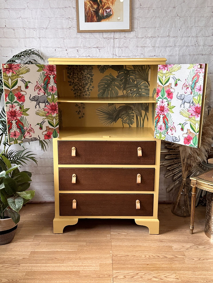 The Hand-painted Tallboy Cabinet stands with its two yellow wooden doors open, revealing a delightful tropical-themed wallpaper inside, adorned with plants and flamingos. The cabinet features three lower drawers, each showcasing brown fronts with stylish leather handles. It is beautifully complemented by surrounding potted plants and a wicker basket. This upcycled linen cupboard offers versatile storage options for the bedroom or kitchen.