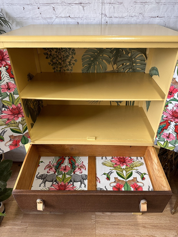 A hand-painted tropical-themed cabinet featuring open yellow shelves and a brown drawer at the bottom, with the inside back and drawer adorned in wallpaper showcasing large leaves, pink flowers, and elephants. The background of the image is also decorated with floral patterns.