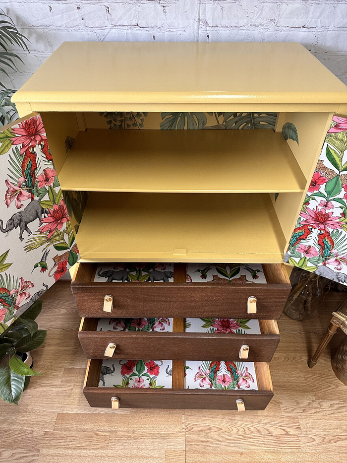 Introducing our Hand-painted Tropical Tallboy! This unique upcycled cabinet features a vibrant yellow top section with two open shelves. Below, you'll find three brown drawers with floral and elephant-patterned interiors. The sides and drawers are adorned with tropical-themed wallpaper, adding an exotic touch to any room. Perfect for bedroom storage, kitchen storage, or clothes storage, this tallboy sits beautifully on a wooden floor surrounded by plants.