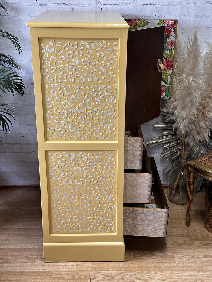 A Hand-painted Tallboy cabinet showcases striking leopard print designs on its side panels and drawer fronts, with one drawer and a cupboard door open to display the floral-patterned interior. The vibrant cabinet stands on a wooden floor, surrounded by potted plants and an elegantly arranged pampas grass display.