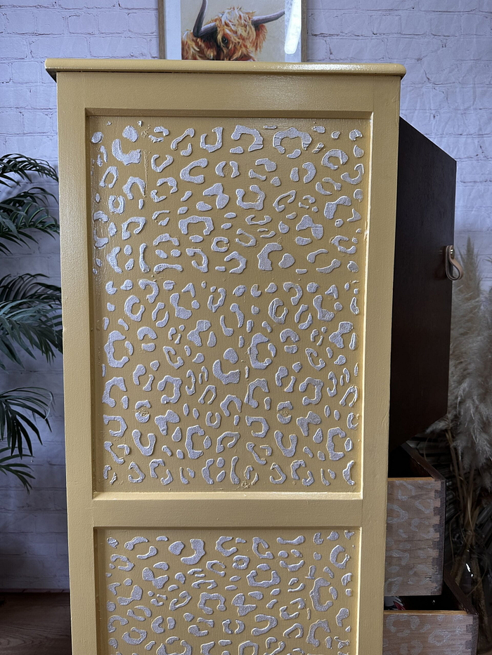 A tropical-themed hand-painted tallboy with leopard print cutouts stands in front of a light-colored brick wall. Above the cabinet, there is a framed picture of an animal on the wall. The scene also includes a potted plant, part of a wooden piece of furniture, and dried pampas grass.