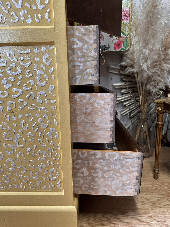 A hand-painted Tallboy and linen cupboard boasts a striking gold and white leopard print design with partially open drawers, set against a backdrop of floral wallpaper and dried pampas grass in the corner. A wooden chair adorned with golden details is also visible to the side.