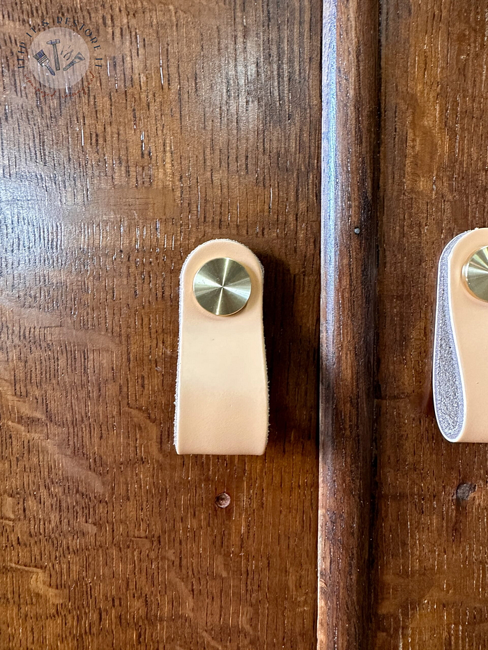 Close-up of a hand-painted, upcycled wooden cabinet featuring a light tan leather pull secured by a round, gold-colored knob. The leather pull is positioned vertically on the cupboard door. The wood has a rich, dark brown finish with visible grain texture.