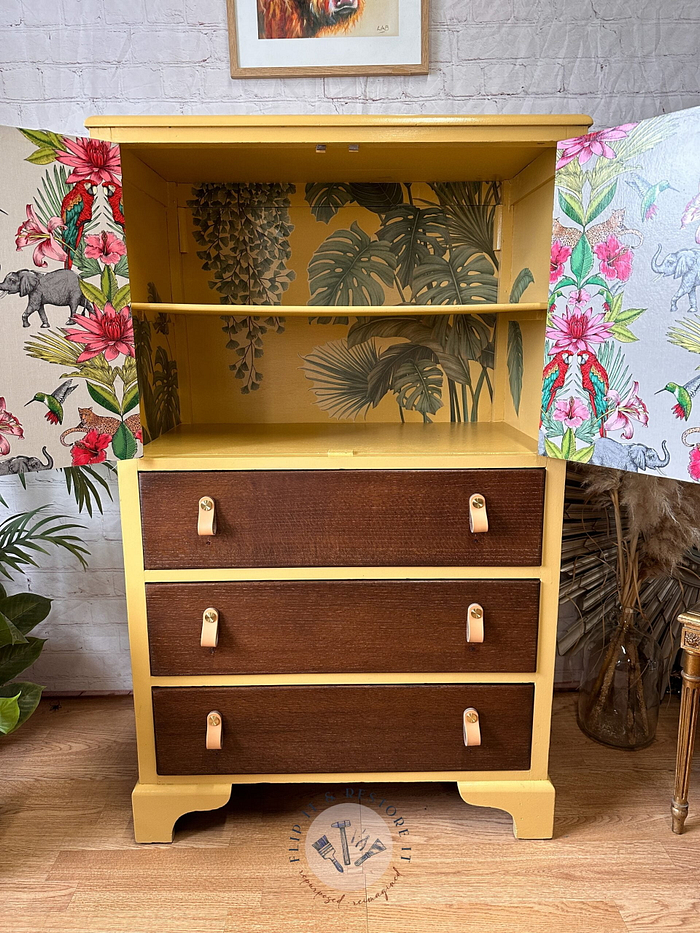 A refurbished yellow "Hand-painted cabinet, Tallboy, Linen Cupboard, Upcycled Cupboard" with three brown drawers featuring leather handles. The cabinet's open doors reveal tropical-patterned wallpaper inside with plants and a few animals. Part of a patterned wall can be seen in the background, along with a potted plant. Perfect for bedroom storage or kitchen storage.