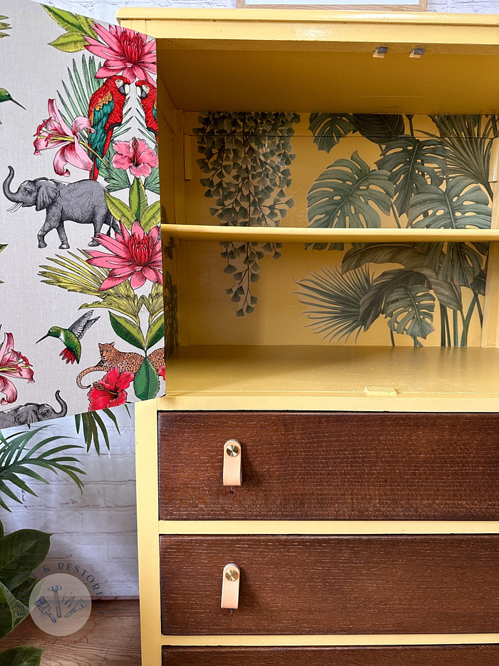 The Hand-painted Tallboy Linen Cupboard presents a vibrant, jungle-themed design. The left side showcases lively wallpaper adorned with a parrot, an elephant, and flowers. The open cabinet reveals shelves decorated with tropical leaves. Below, three wooden drawers feature light brown handles, adding to its unique charm.