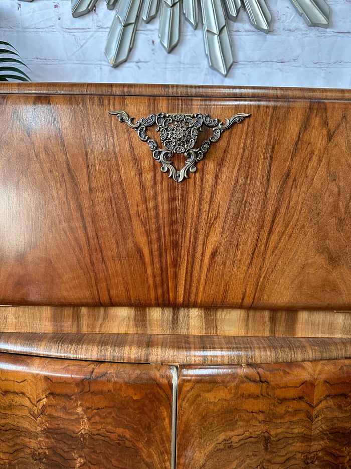 Close-up of the Rivington Cocktail Cabinet, featuring vintage Art Deco design with intricate detailing at the top center. The rich walnut and polished green and gold finish are visible, with decorative monstera leaves adding an elegant touch. The background includes a decorative, radial mirror reflecting its retro charm.