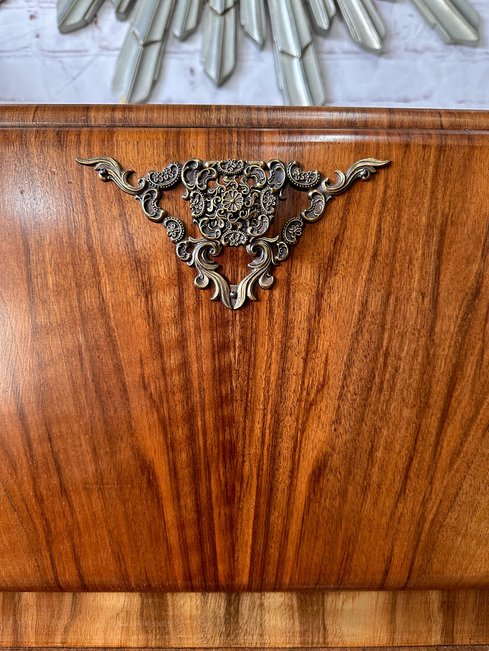 A close-up of the Rivington Cocktail Cabinet, an ornate wooden piece from the 1950s with a vintage art deco aesthetic. The cabinet, made from walnut and featuring green and gold accents, showcases a decorative metal embellishment in the center with intricate scrolling patterns and a floral design. Above, part of a sunburst mirror accented by metal rays is visible.