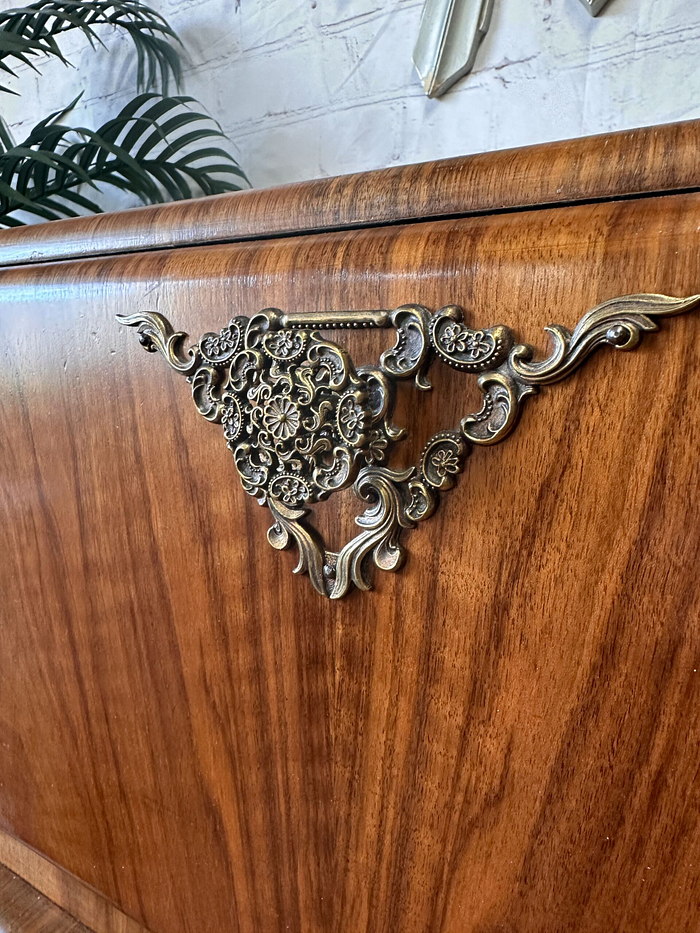 Close-up of an ornate, brass corner embellishment on a wooden piece of furniture. The intricate design features floral and scroll elements. A green leafy Monstera plant is partially visible in the background, and there are white brick wall details behind the Rivington Cocktail Cabinet, a vintage drinks bar with an art deco style from the 1950s, finished in green and gold walnut.