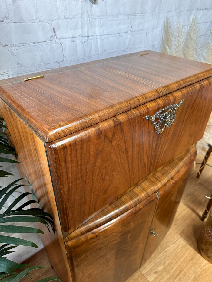 The Rivington Cocktail Cabinet, a vintage drinks bar from the 1950s featuring an Art Deco design, stands proudly on a wooden floor. This retro bar, crafted from walnut with a green and gold finish and adorned with monstera leaves motifs, boasts rounded edges and two closed compartments. Positioned against a white brick wall, it is complemented by the presence of green foliage on the left side.