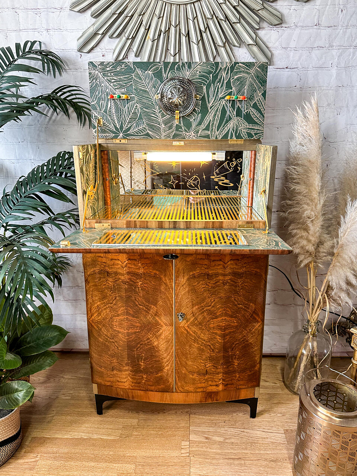 A stylish vintage Rivington Cocktail Cabinet, featuring detailed wood grain patterns and a glass display with a lit interior, stands against a white brick wall adorned with botanical wallpaper. The green and gold Art Deco piece from the 1950s is accentuated by ornate decor and surrounded by plants and pampas grass in vases, enhancing the aesthetic appeal.