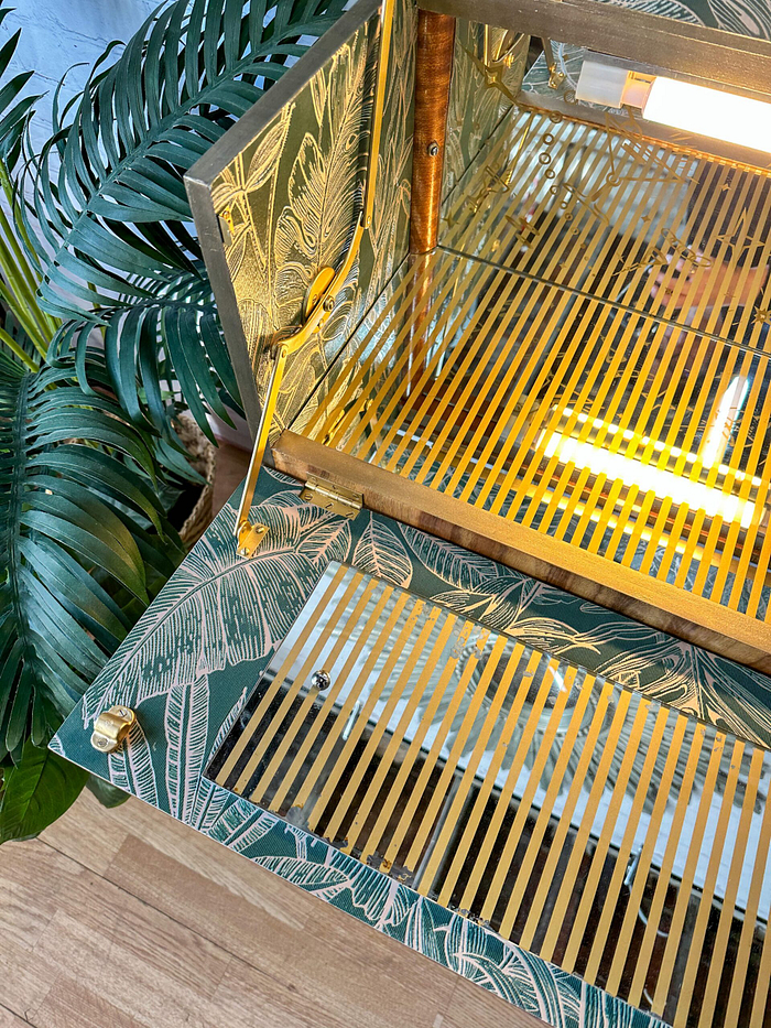 The Rivington Cocktail Cabinet with its tropical monstera leaf print interior and gold-striped glass shelves is open. Warm lighting inside the cabinet accentuates its design. A large green plant with broad leaves is partially visible beside the cabinet on a wooden floor.