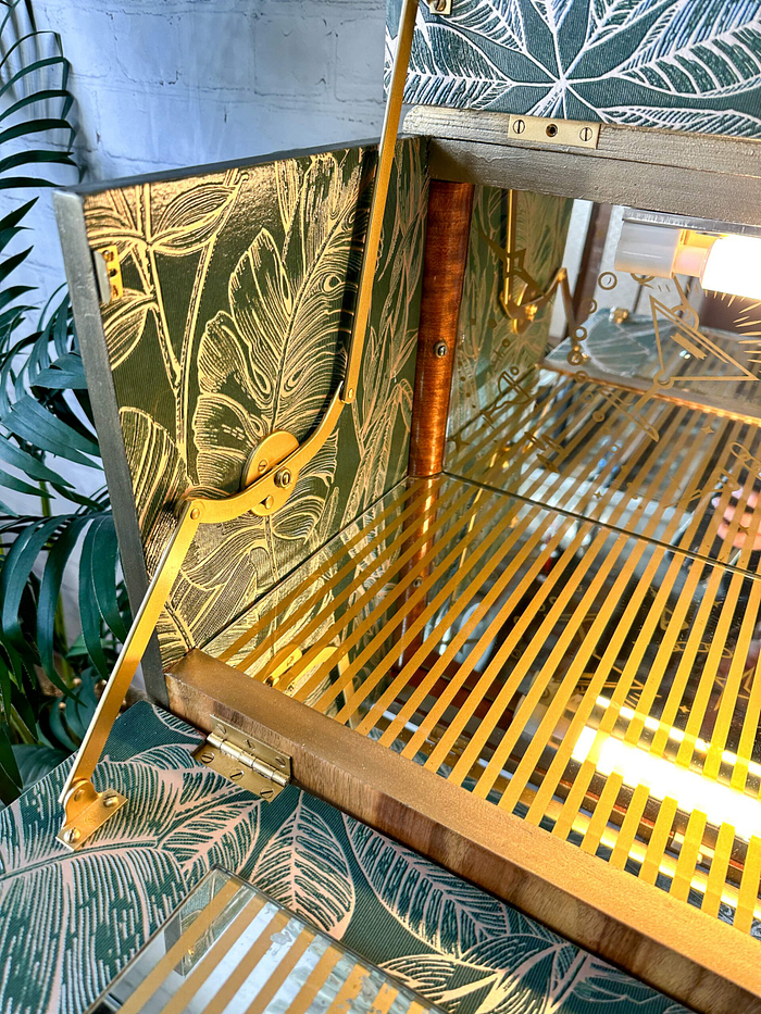 A close-up of the meticulously crafted interior of the Rivington Cocktail Cabinet, featuring vibrant monstera leaf-patterned wallpaper. The vintage drinks bar includes elegant gold-colored hinges and slats, complemented by reflective mirrored surfaces. Green foliage and a white painted brick wall can be seen in the background, enhancing its retro charm.