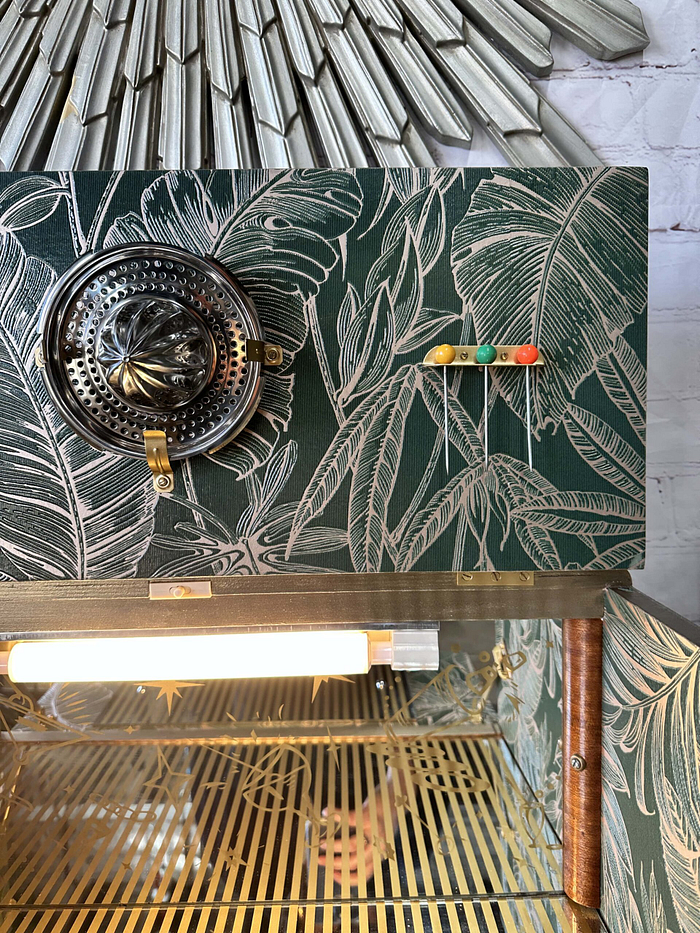 The Rivington Cocktail Cabinet showcases a striking decorative wall with a vintage tropical leaf pattern in silver on a dark green backdrop. A silver ventilation fan is positioned on the left, while four small colored balls are neatly aligned on the right. Beneath it, a lit display shelf with an elegant gold-striped metal surface completes the iconic Art Deco look of this 1950s-style retro bar unit.