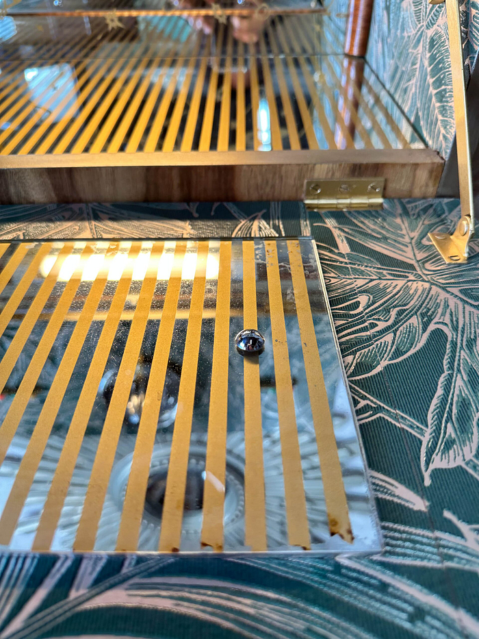 A close-up of the Rivington Cocktail Cabinet shows off its glass lid adorned with gold vertical stripes. The cabinet is open, showcasing a mirrored interior. On the mirrored surface rests a reflective silver bead. In the background, a green pattern with white monstera leaves enhances the vintage Art Deco aesthetic of this 1950's retro drinks unit.