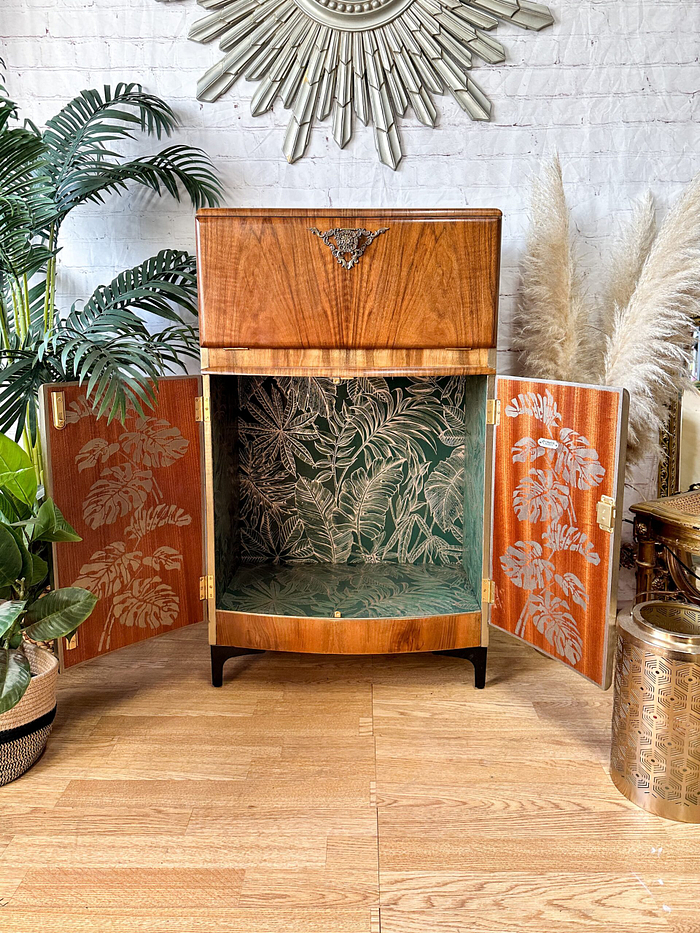 The Rivington Cocktail Cabinet, a vintage walnut drinks bar from the 1950s, is shown with its doors open to reveal an interior adorned with green and white monstera leaf wallpaper. This retro Art Deco unit features elegant brass accents and is complemented by surrounding plants and a fur rug. The setting includes a white brick wall and a decorative sunburst mirror in the backdrop.