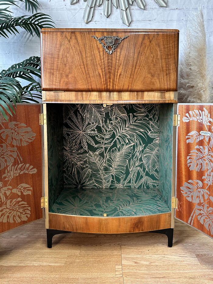 The Rivington Cocktail Cabinet, a vintage walnut drinks unit from the 1950s, stands open to reveal an interior lined with green wallpaper adorned with a leafy Monstera pattern. The outer doors also exhibit a botanical design. Partially visible in the background are indoor plants and a decorative wall mirror, enhancing its retro Art Deco charm.