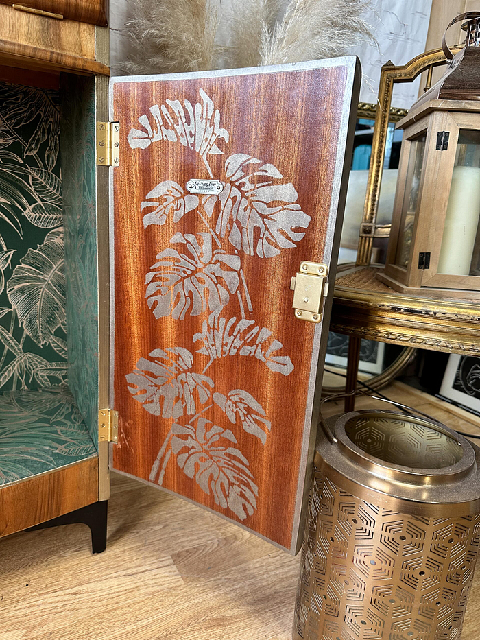 An open door of the Rivington Cocktail Cabinet reveals an intricate design of Monstera leaves. Adjacent to it, a golden decorative lantern featuring geometric cutouts with a white candle is set on the walnut wooden floor. The top of the cabinet is adorned with dry grass decorations, adding a touch of natural elegance.