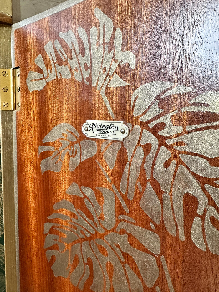 A wooden Rivington Cocktail Cabinet showcases an intricately painted design of tropical Monstera leaves. A small metallic label on the cabinet reads "Rivington Product, London." The unit features a brass hinge visible on the left side.