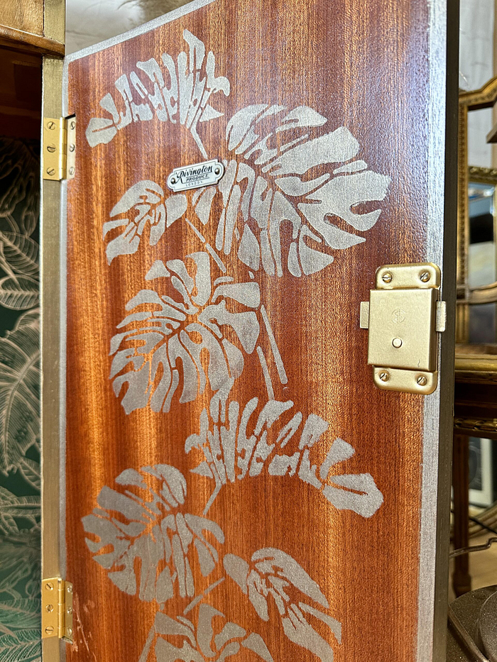 A close-up of the Rivington Cocktail Cabinet showcases its wooden exterior with metallic hinges and a latch, adorned with an intricate silver leaf pattern. A small metal label on the top plank reads "Handcrafted." In the background, there's a glimpse of another cabinet featuring botanical patterns.