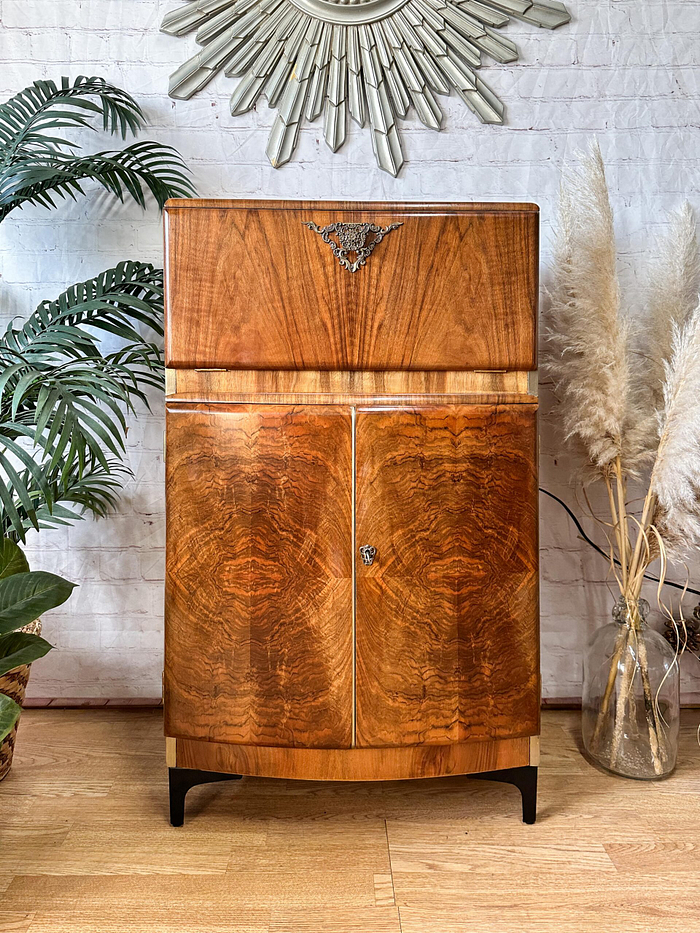 The Rivington Cocktail Cabinet, an Art Deco vintage drinks bar from the 1950s, features a stunning walnut finish with intricate monstera leaf patterns in green and gold. This retro bar unit boasts a decorative metal element at the top and double doors at the bottom. It is elegantly placed against a white brick wall, framed by lush green plants on the left and dried pampas grass in a vase on the right.