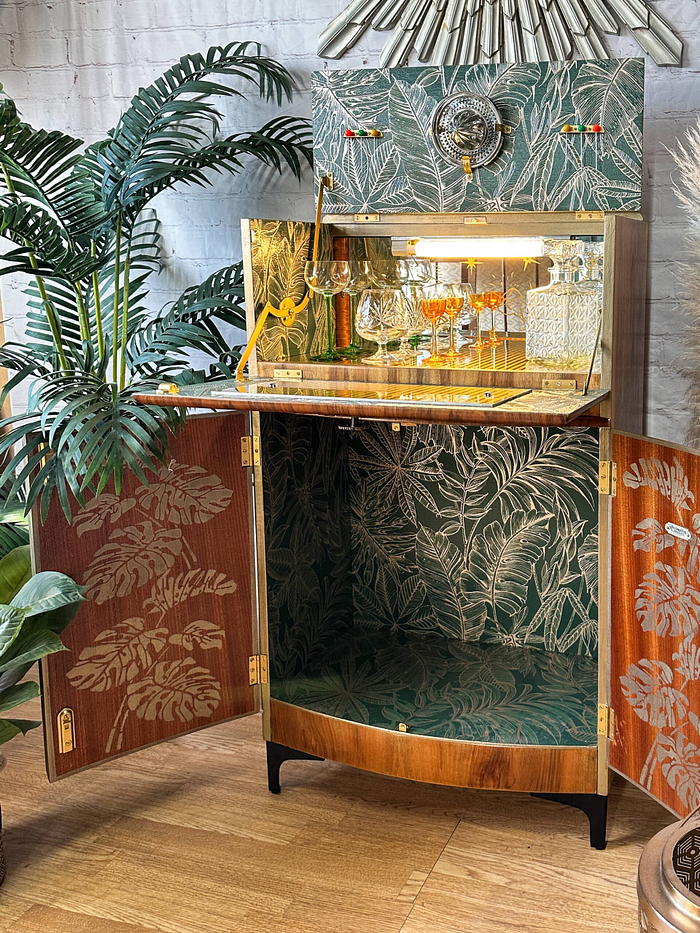 The Rivington Cocktail Cabinet is open, showcasing a mirrored back adorned with Monstera leaf patterns and glass shelves stocked with glasses and bottles. The vintage walnut cabinet features stylish green and gold Art Deco designs on its doors, blending seamlessly with the lush greenery surrounding it on the wooden floor.